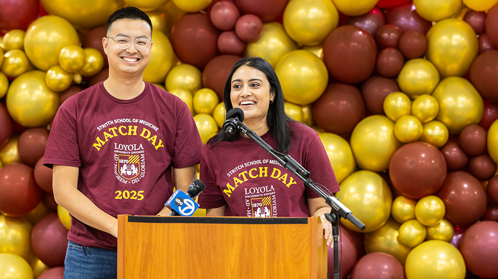 Two students at a podium with wall of maroon and gold balloons behind them.